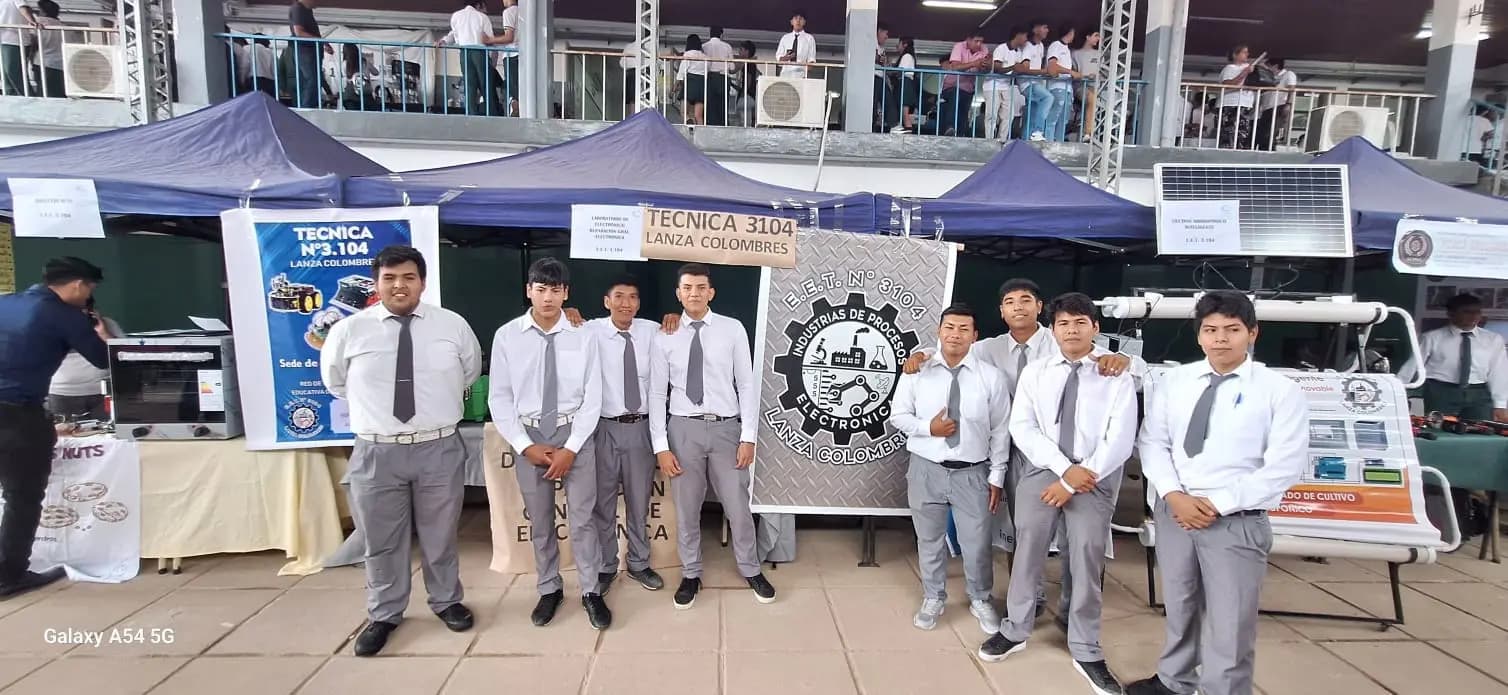Alumnos de la escuela frente al stand de la Técnica 3104 en una feria, con sus proyectos y el banner de Industria de Procesos y Electrónica