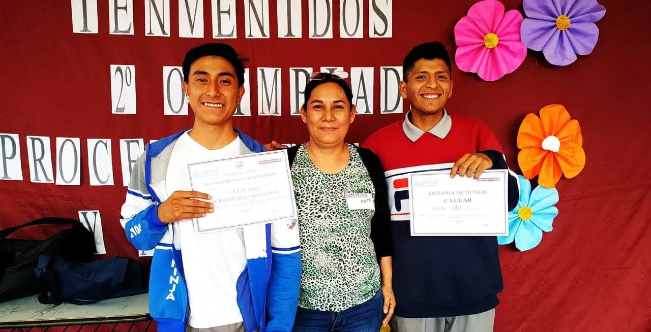 Dos alumnos con el diploma de honor de primer lugar y el reconocimiento de la escuela, junto a su docente, frente al cartel de la olimpíada